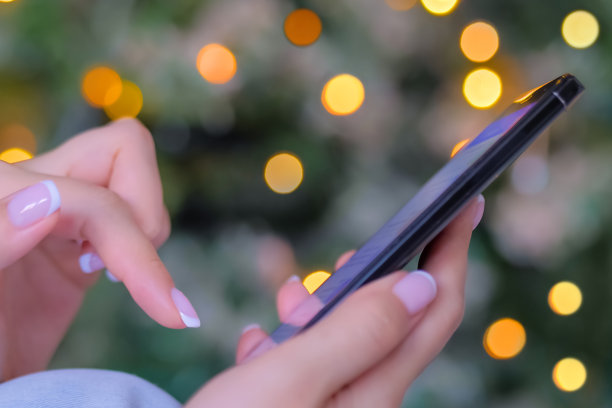 近侧视图-女人使用智能手机，圣诞灯光背景图片下载