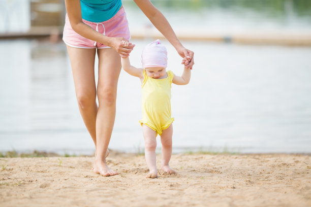 年轻的母亲帮助小女孩在海滩上迈出第一步。可爱的，和平的漫步在夏日的沙滩上。前视图。素材图片