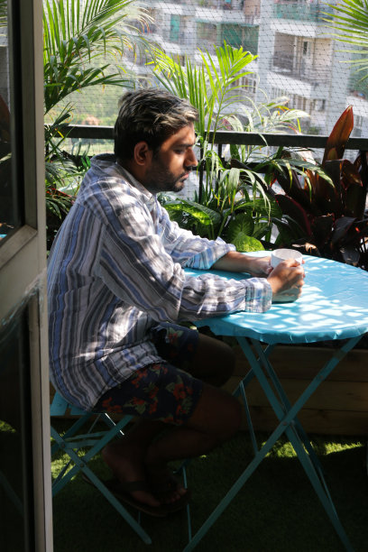 一个男人坐在阳台露台上的蓝色圆桌旁，在阳光下喝着一杯热咖啡，从走廊厨房门口观看图片下载