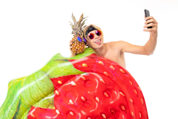 年轻的白人金发男子站在泳衣与大橡胶沙滩床垫做自拍与菠萝孤立在白色的背景图片下载