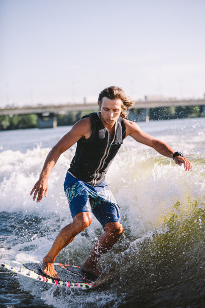漂亮的金发碧眼的浪头，穿着背心在船上沿着湖面的波浪。健壮的男运动员在夏天在河上冲浪醒来。Wakesurf水上运动。在波冲浪。一个人在冲浪板上冲浪图片下载