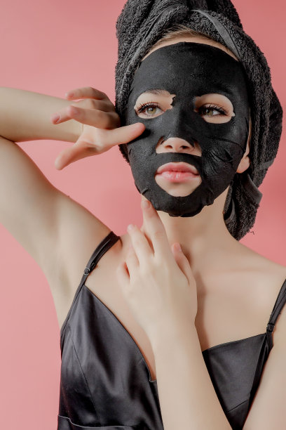 年轻女子将黑色化妆布面膜涂在粉色背景上。面部脱皮用炭面膜、spa美容、护肤、美容。近距离图片下载