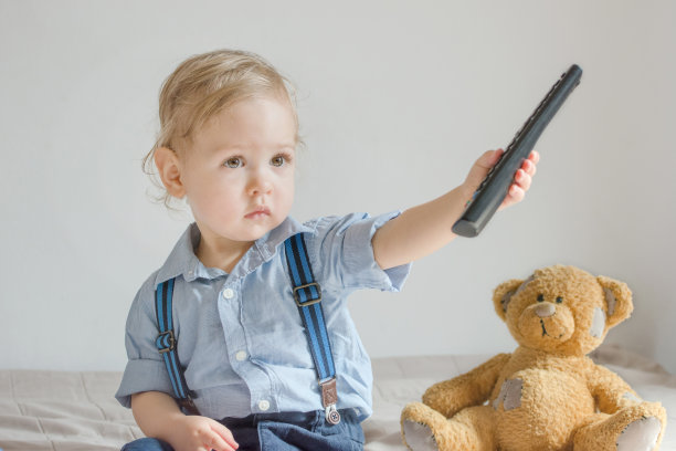 可爱的小男孩玩着遥控器看电视，坐在沙发上和他的泰迪熊，在家里图片下载