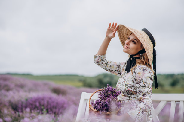 年轻美丽的金发女子穿着裙子，戴着草帽，带着花篮，坐在普罗旺斯风格的白色长凳上，面带微笑。软选择聚焦。图片下载