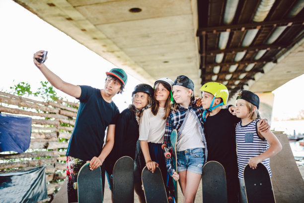 一群玩滑板的朋友在滑板公园用手机自拍。一大群带着滑板的孩子在滑板公园的坡道上拍摄回忆照片。一群孩子在智能手机上合影图片下载