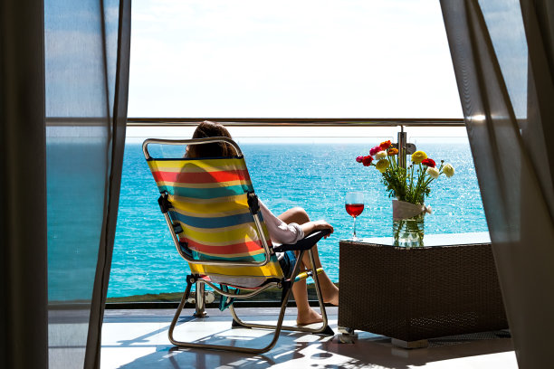 长发女子坐在户外阳台的躺椅上，带着鲜花和酒杯，看着阳光明媚的海边景色与蓝天，后视镜。图片下载