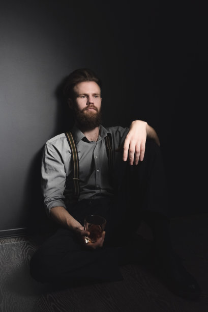 一个留着胡子的英俊年轻人手里拿着一杯酒坐在一间黑屋子里。休息和放松，酗酒和孤独的概念图片下载