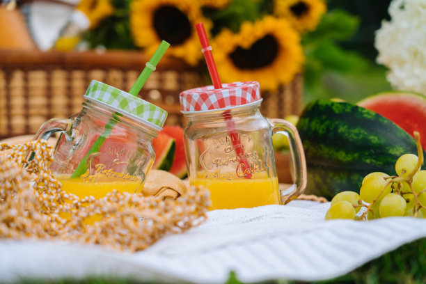 夏日野餐的概念在阳光明媚的日子有西瓜，水果，绣球花和向日葵花。草地上的野餐篮子，上面放着食物和提神的夏日饮料，上面盖着白色的针织毯子。有选择性的重点图片下载