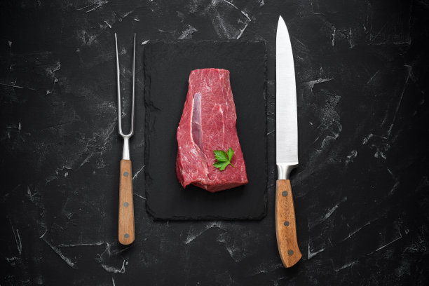 新鲜的牛排与刀叉在黑色的石头背景。生肉角。俯视图，平放，复制空间。图片下载