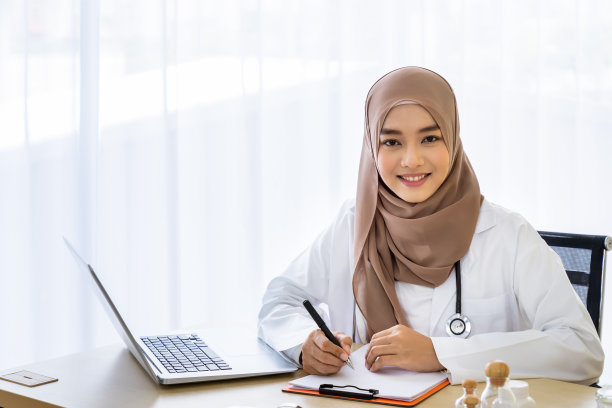 自信的穆斯林女医生坐在诊所医院办公室的办公桌前，在笔记本上写作，看着镜头图片下载