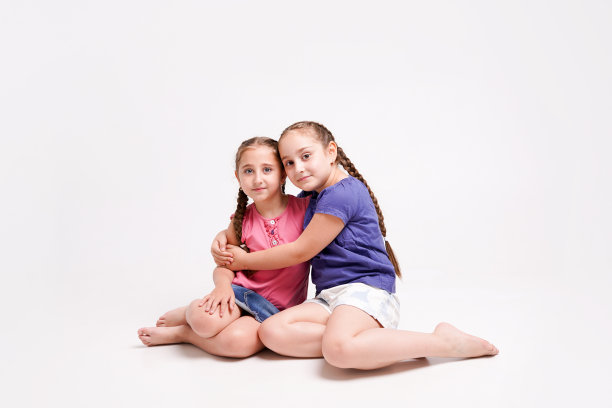 有趣的女孩，姐妹们坐在地板上跪着，拥抱在白色的背景图片下载