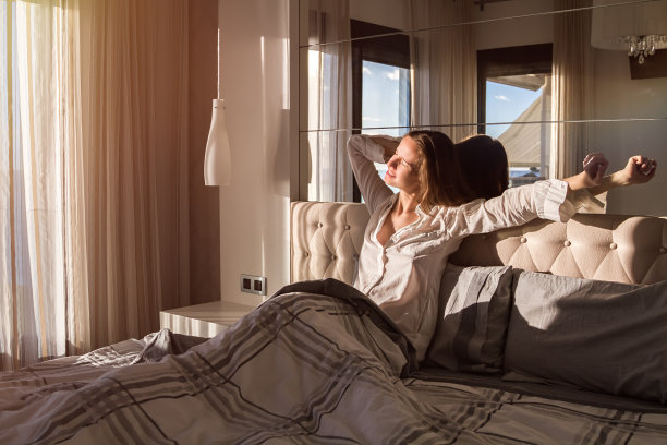 一个美丽的年轻长发女人的肖像早晨从第一缕阳光和伸展她的手臂，坐在床上在一个现代的卧室。图片下载