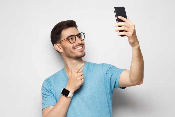 一名身穿白色t恤、手腕上戴着手表的年轻人，拿着手机自拍，孤立在灰色背景中图片下载