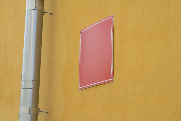 落水管旁边的黄色墙壁上空白的红色塑料牌匾。与墙成一个角度的有放置文字的地方的盘子。图片下载