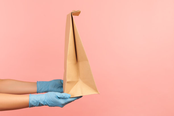 侧面特写的人的手在蓝色外科手套持有和牛皮纸包装的食物。安全食品配送理念。图片下载