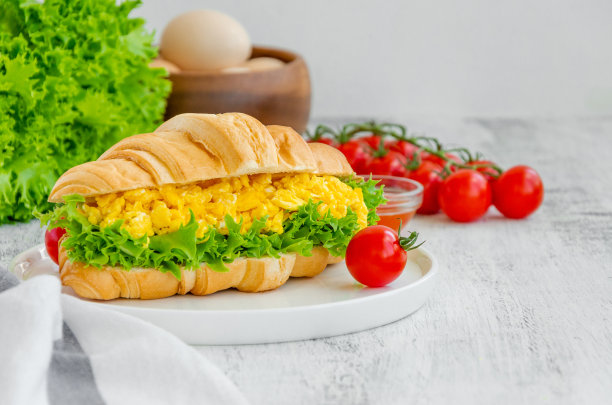 牛角面包，炒鸡蛋，生菜和樱桃番茄放在一个白色的盘子里，食材在木制的背景上烹饪。水平,复制空间。图片下载