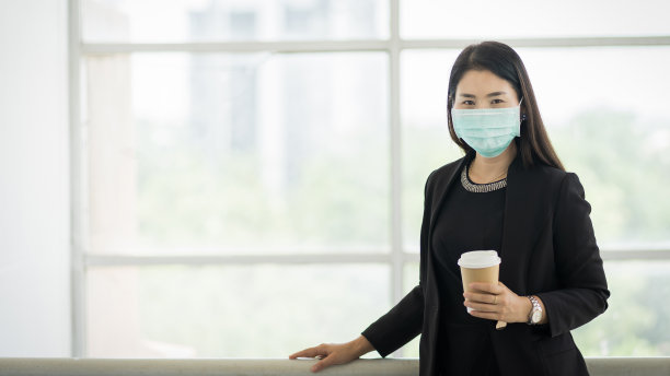 一个商务女性的肖像，穿着商务套装戴着面具，在公司大楼早上咖啡杯准备工作。一名戴口罩的中年妇女，以保护COVID-19大流行。库存图片图片下载