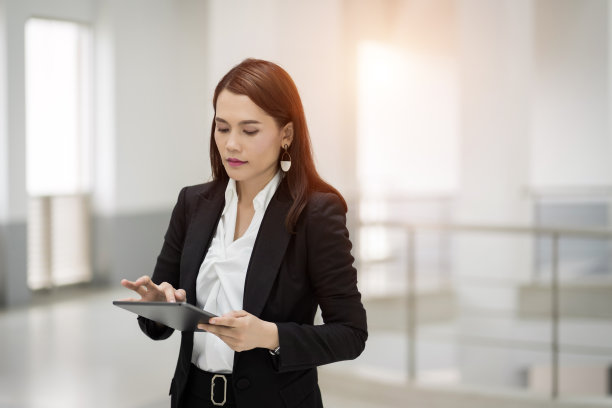 这是一位年轻开朗的女商人在休息期间在办公室前用平板电脑浏览社交网络的照片。站在办公大楼里的亚洲女商人。库存图片图片下载