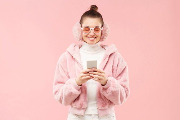年轻的学生女孩穿着冬衣，戴着耳罩和眼镜，手里拿着手机，和朋友们交换短信，在粉红色的背景下孤立无援图片下载