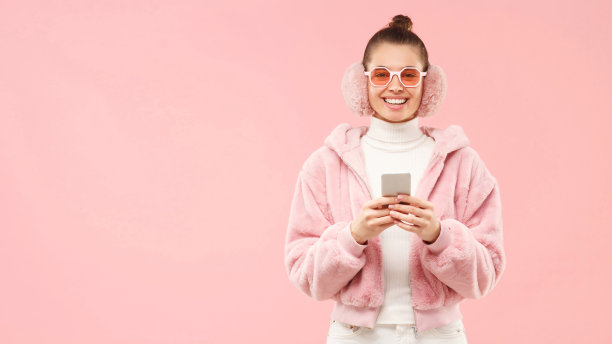 一个年轻快乐的女孩穿着冬天的外套，戴着耳罩和时髦的眼镜，手里拿着智能手机，孤立在粉红色的背景和左边的拷贝空间图片下载