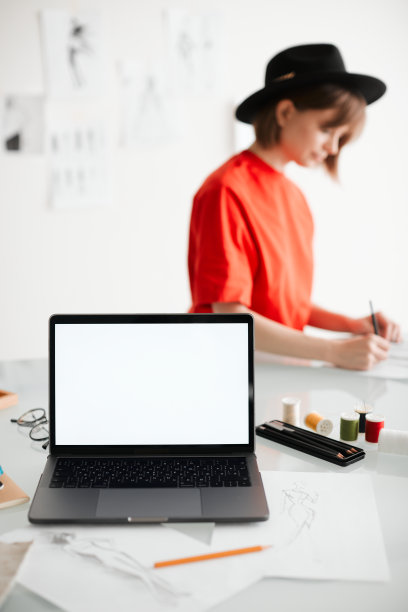 年轻女孩时装设计师在帽子和笔记本电脑空屏幕在工作室。图片为广告，教育课程或服装图片下载