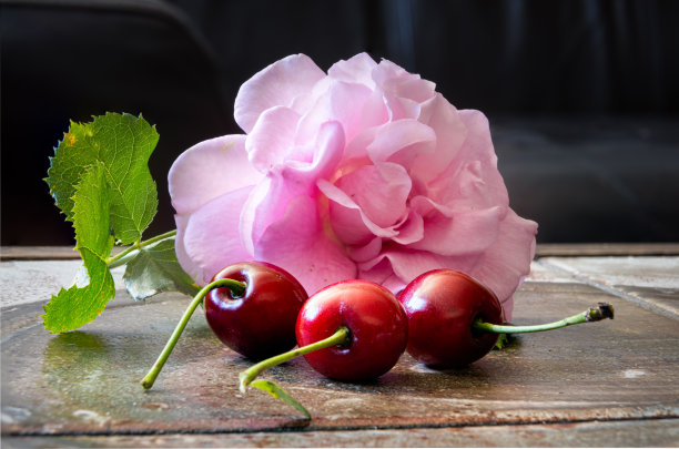 这是三颗红樱桃的特写镜头，上面有粉红色的玫瑰和棕色表面上的绿叶。微距照片与黑暗的背景。插图图片下载