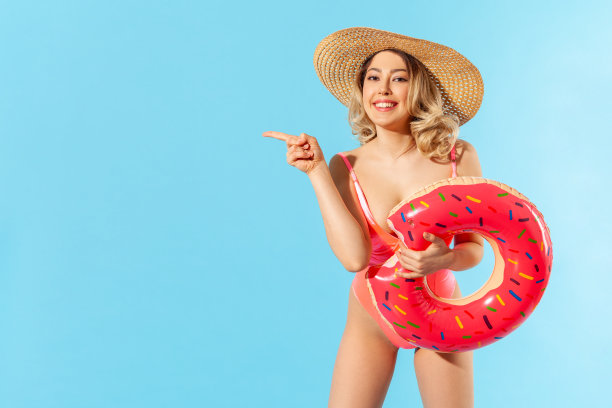 看,广告!快乐的女人穿着泳衣和太阳帽拿着橡胶圈，指着拷贝空间，空白墙图片下载