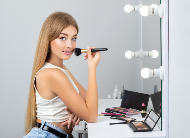 年轻漂亮的金发女郎，皮肤干净清新，长长的金发拿着刷子，在有灯的镜子前化妆。她旁边有很多眼影和粉。化妆的概念图片下载