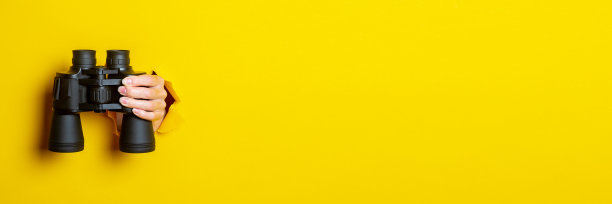 女性手持黄色背景上的黑色双筒望远镜。通过双筒望远镜，旅行，寻找和搜索的概念。横幅。图片下载