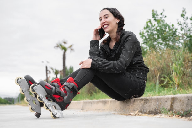 快乐的女人在滑旱冰时停下来，坐在街上玩手机。穿着旱冰鞋，打电话的漂亮都市女孩。都市现代女孩。高质量的照片图片下载
