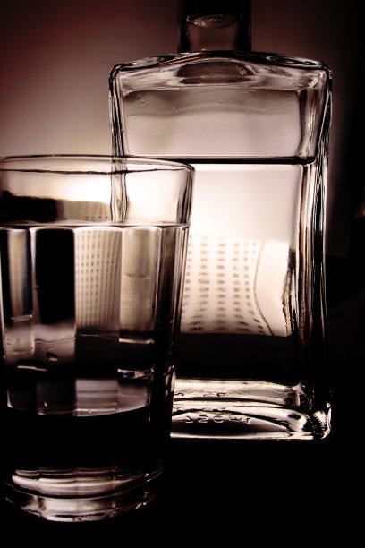 一个长方形的瓶子和一个透明的玻璃杯，装满了无色的液体，在半影的背景上。图片下载