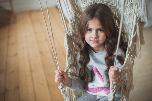 一个穿着灰色睡衣的漂亮的黑发女孩在秋千上放松。可爱的黑发女孩在家里的编织椅摇摆。看着摄像机。特写,柔软的焦点。图片下载