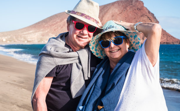 老夫妇在沙滩上拥抱，在一个有风的日子在沙滩上散步。两个戴有色帽子和太阳镜的人。背景是红色的山。天空和蓝色的海洋图片下载