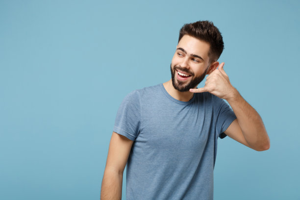 年轻开朗的有吸引力的男人在休闲衣服摆姿势孤立在工作室的蓝色背景。人们生活方式的概念。模拟拷贝空间。做着手机手势，好像在说给我回电话，然后看向一边。图片下载