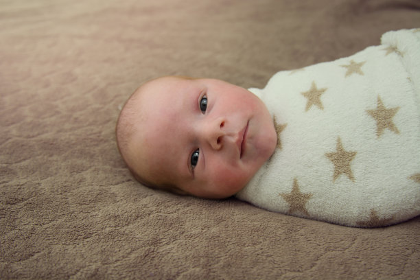 一个裹着白色襁褓的可爱新生儿躺在棕色的毯子上看着镜头。美丽的小男孩的肖像7天，一个星期。Copyspace图片下载