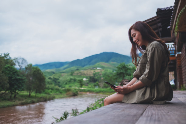 一个美丽的亚洲女人在使用手机，而坐在河边与山和自然图片下载