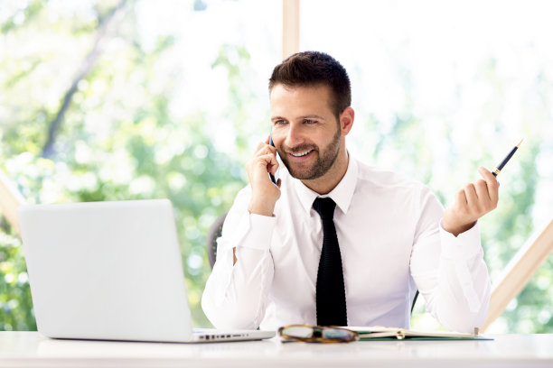 自信的商人坐在办公桌旁用手机和笔记本电脑与人交谈图片下载