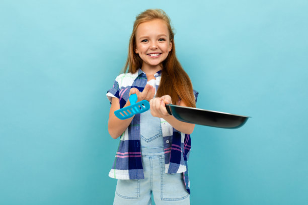 一个面带微笑的欧洲女孩，手拿厨具站在浅蓝色的墙上图片下载