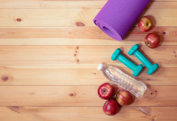 紫色的瑜伽垫，绿色的哑铃，一瓶水和木制背景上的红苹果。图片下载