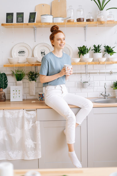 美丽的红发年轻女子拿着一杯热咖啡，而站在厨房。图片下载