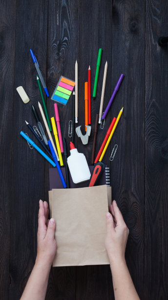 学校背景，学习用品，写工具在牛皮纸包上，在一个深色的木制背景，旁边的女人的手图片下载