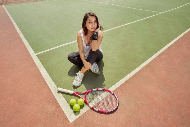 女网球运动员，穿着休闲服，球拍搁在网球场上。高角度的漂亮女人在时尚的白色衣服坐在网球场上与球和球拍周围。图片下载