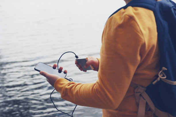 这家伙手里拿着一个便携式充电器和一个智能手机。一个背着背包的男子站在湖边，用电源银行给电话充电。关于旅游主题的概念。图片下载