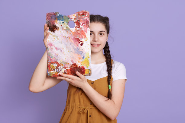 图片的温柔年轻的女士拿着照片在手中，覆盖一半的脸，穿着衣服，站在丁香的背景，可爱的艺术家展示她的工作孤立。图片下载