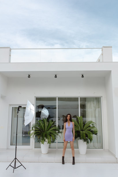 苗条迷人的时尚模特穿着紧身的淡紫色连衣裙，在棕榈树、高科技建筑和摄影设备的背景下摆姿势。图片下载