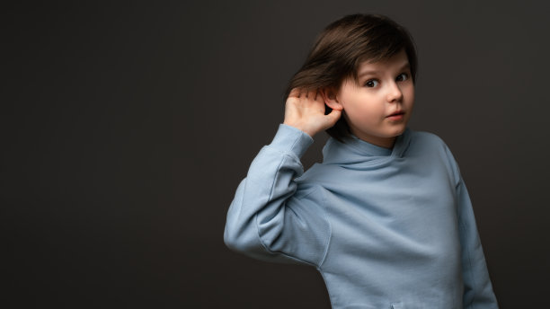 好奇的10-12岁的小男孩，手放在耳朵上，试图偷听私人谈话，穿着休闲衣服，在灰色的演播室背景下摆姿势图片下载