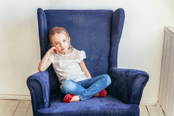 甜美的小女孩在牛仔裤和白色t恤坐在现代舒适的蓝色椅子放松在白色明亮的客厅在家里室内。儿童学龄儿童青年放松观念图片下载