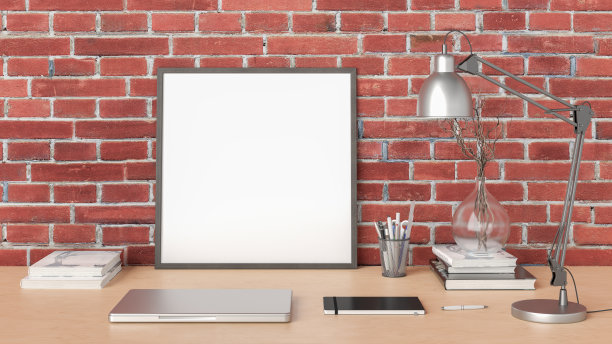 方形海报框架模型在家庭工作室工作空间的木桌上与红砖墙。正面视图，剪贴路径周围的海报图片。图片下载