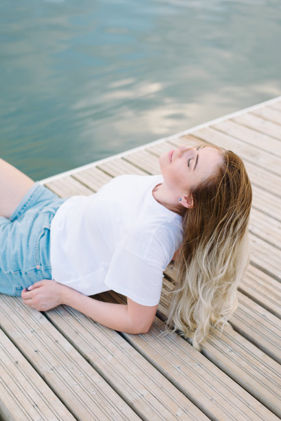 一个穿着白色t恤的女人坐在水边的木制码头上。布隆代,微笑图片下载