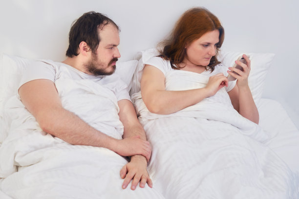 丈夫躺在白色的床上看着妻子打电话。一个男人偷看一个女人的电话。夫妻二人在隔离期间待在家里。图片下载